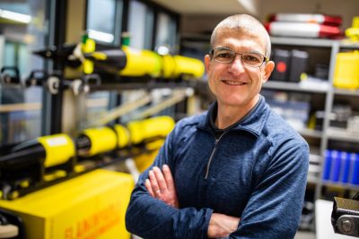 A man with thinning salt and pepper hair, glasses, and a blue shirt poses for a photograph in a lab with robotic submarines.