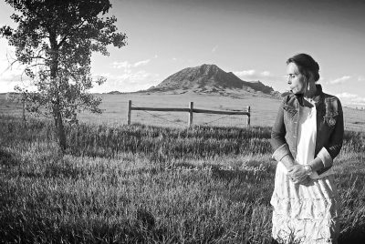 In this black and white image, author and activist Winona LaDuke - a Native American woman with dark hair pulled up in a bun wearing a frilly dress under a denim jacket - looks away from the camera, down at the ground. She stands in a friend with a tree behind her and a large mountain in the distance.