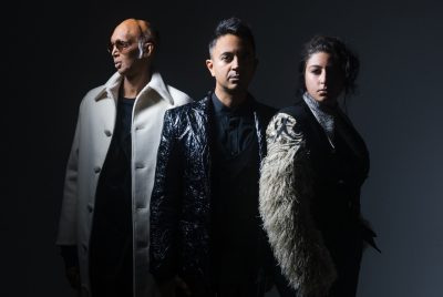 From left, Shahzad Ismaily, a bald brown man with white sideburns, round sunglasses, and a long white peacoat over a dark shirt; Vijay Iyer, a young brown man with medium length dark brown hair in a fade on the sides, wearing a black and silver brocade blazer over a black button down shirt, and Arooj Aftab, a young brown woman with dark brown hair pulled into a messy updo, wearing a black jacket with fluffy white sleeves. They stand in front of a dark grey background.
