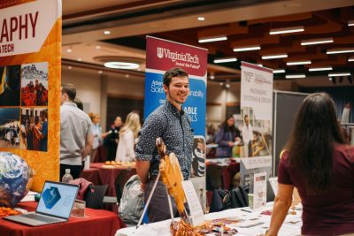 A man smiles from behind a table leaden with materials, in front of banners advertising Virginia tech majors, at the majors and minors fair.