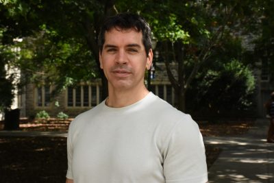 A man with dark hair and wearing a white t-shirt stands near some trees.