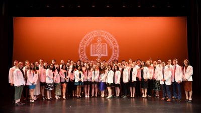 A group of 51 first-year students from the Virginia Tech Carilion School of Medicine stand together on stage wearing their white coats.