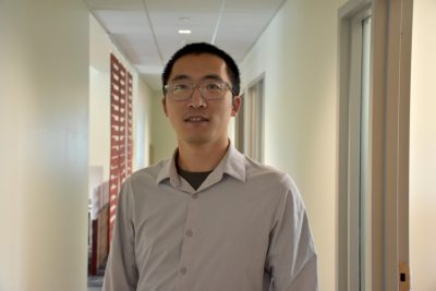 A young professor with dark hair and a beige dress shirt stands in a hallway.