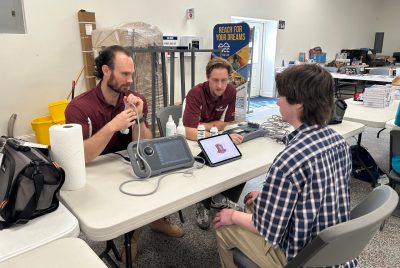 Two men wearing VTCSOM shirts showing a younger man a portable ultrasound machine