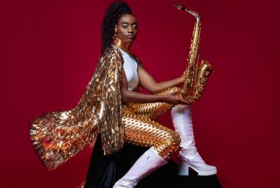 Jazz saxophonist Lakecia Benjamin, a young Black woman with twists pulled up into a side ponytail, wears a gold cape and pants and white shirt and boots. She sits in front of a red background and holds her saxophone up and away from her body, gazing unconcernedly towards the camera.