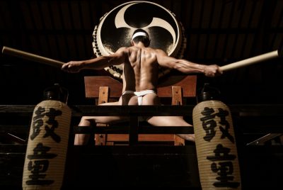 A Japanese drummer wears a white headband and a white fundoshi and extends both arms out side to side, a drum stick in each hand and back muscles all tensed. In front of him is an enormous drum.