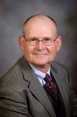 Man in gray suit with a blue dress shirt and tie wearing glasses.
