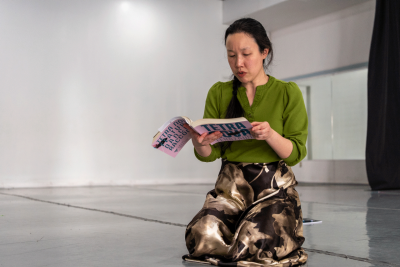 A woman kneels on the floor while reading her book, 'Tetra Nova.' She wears a green shirt and brown flowered shirt.