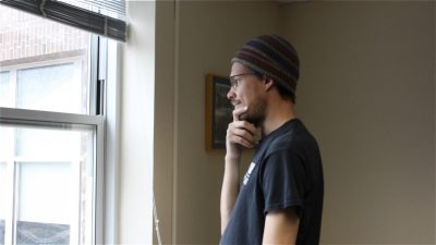 Greene wears a knitted cap and a black t-shirt and glasses while standing and looking out the window of a room in Shanks Hall.