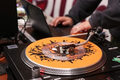 In the background, a set of hands types on a laptop. In the foreground is a record player. 