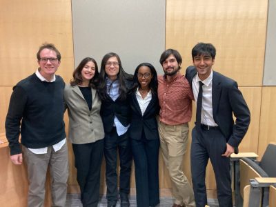 A group of six students, dressed in formal clothing, stand in front of wooden paneling, smiling.