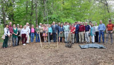 A large group of people stand around a young oak tree planted near the Duck Pond.