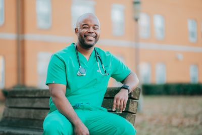 Greg Echols posing for a photo sitting on a bench wearing green scrubs and a stethoscope around his neck