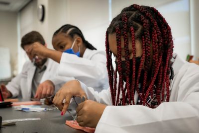 Two young female students in white coats practicing suturing using training materials.  Male in whiute coat observing.,