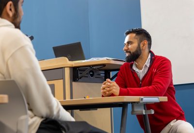 A teacher wearing a bright red sweater kneeling at a desk to talk to a student