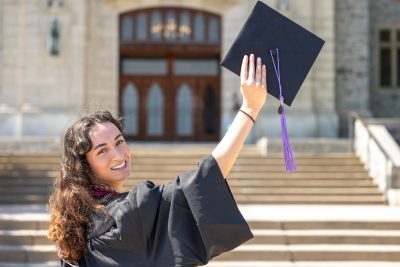 A young woman wearing a black graduation gown stands outside in front of Burruss Hall at Virginia Tech. She is smiling over her shoulder at the camera while holding her graduation cap high in the air with her right hand. The cap has a purple tassel hanging from it. The background shows the stone exterior and large wooden doors of Burruss Hall, with a set of wide steps leading up to the entrance. The weather is sunny and bright, casting clear shadows on the steps.
