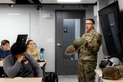 An adult soldier in battle dress uniform holds two helmets of different eras while talking to seated students who look intent.