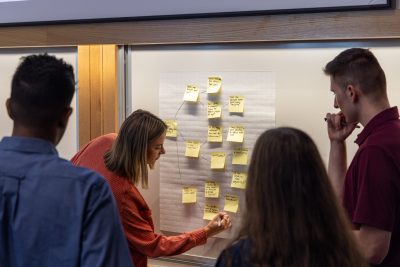 A student writes on a large pad attached to a whiteboard while other students watch